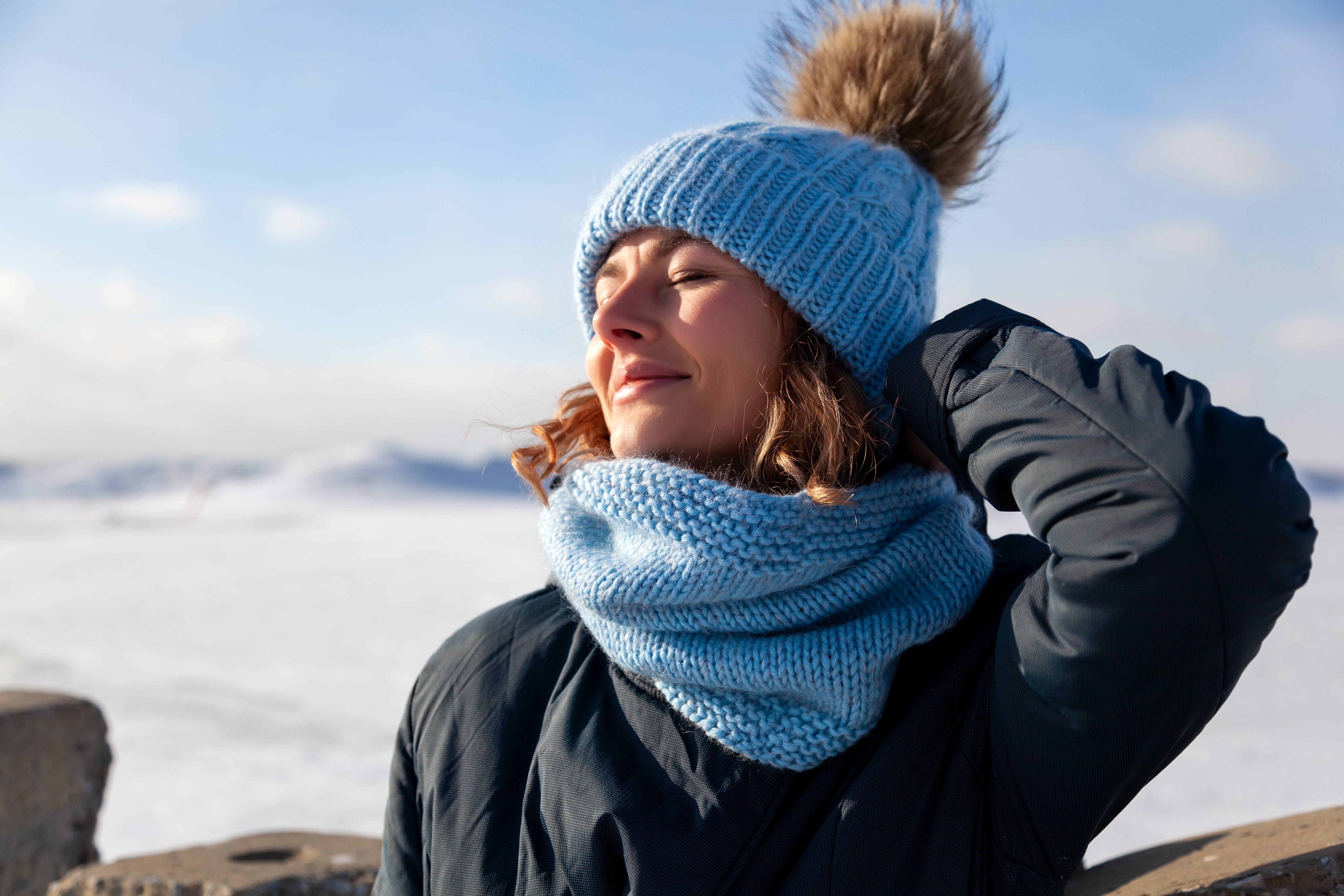 Eine Frau mit blauer Mütze und blauem Strickschal genießt mit geschlossenen Augen eine herrliche Winterlandschaft im Hintergrund